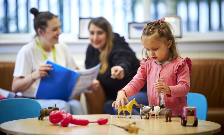 A child standing playing with toy animals on a table. Two adults sitting behind the child talking to each other.