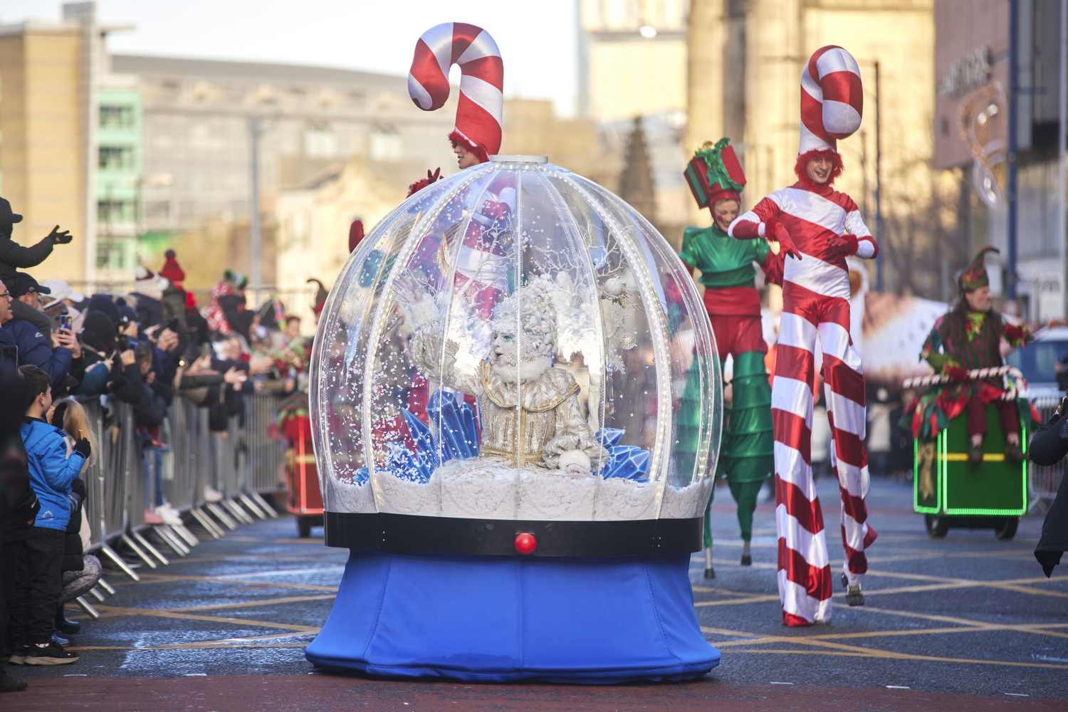 A giant snow globe with a snow queen inside pictured with candy cane stilt walkers either side parading along a street