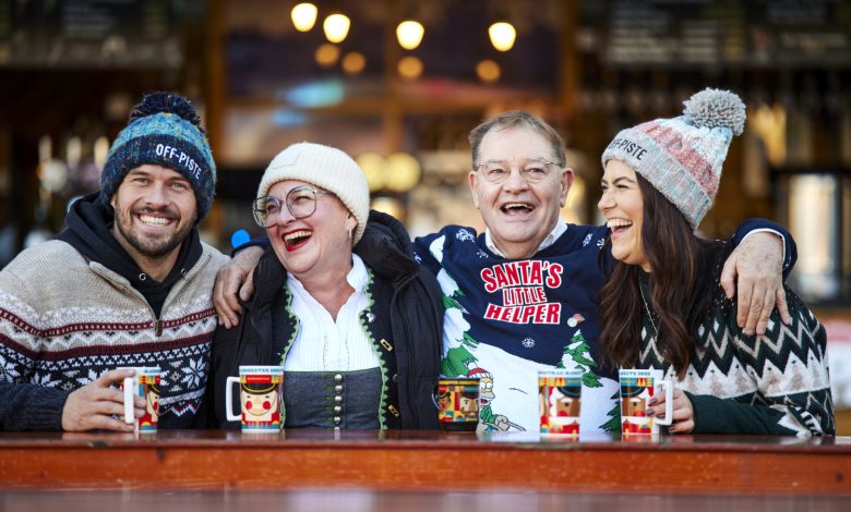 Four people laughing, arms around each other, holding mugs in their hands