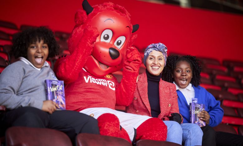 Two children and an adult sit with a bright red adult-sized mascot on the seats of a football terrace