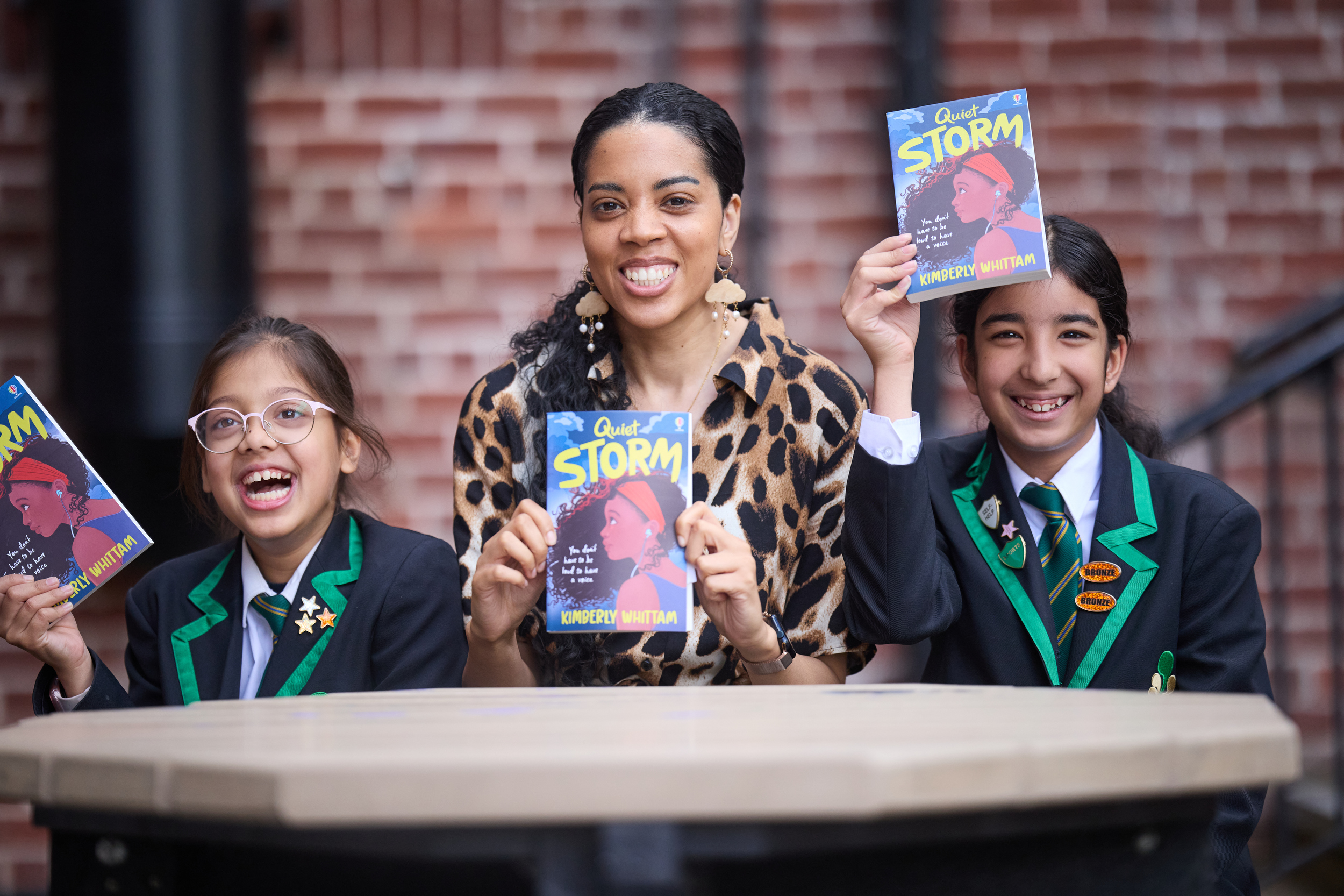 A woman sits inbetween two young school pupils wearing black blazers with green edging.  They are each holding a copy of the same book and are all smiling.