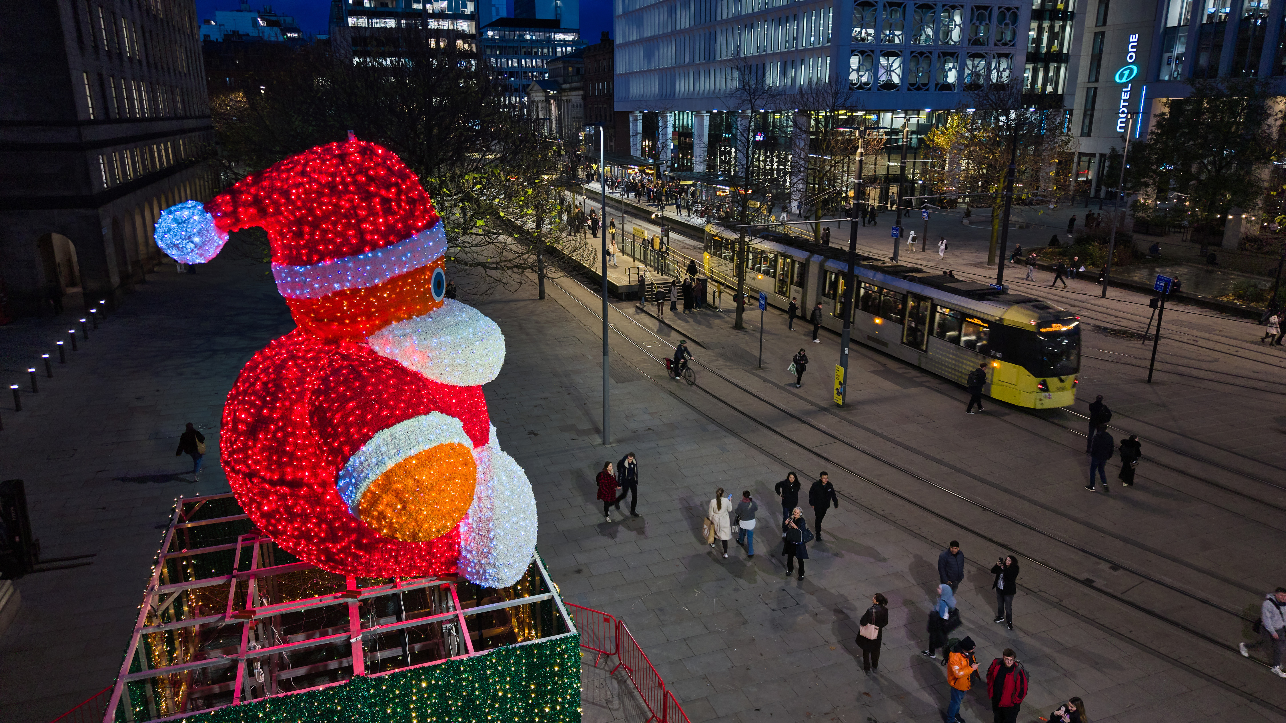 Night time view from the skies of street scene with yellow trams running across St Peters Square in Manchester and a bright red Santa Claus figure sitting on top of a huge present both lit up with sparkly lights